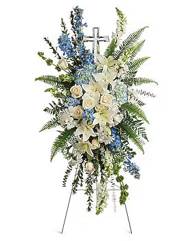 A large floral funeral arrangement on a metal easel stand, featuring white and blue flowers with green foliage and a silver cross at the top, set against a plain white background.