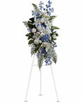 A large floral wreath or floral arrangement displayed on a white easel stand against a plain white background. The bouquet features white lilies, blue flowers, green foliage, and fern-like leaves.