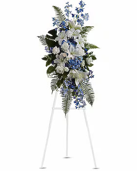 A large floral wreath or floral arrangement displayed on a white easel stand against a plain white background. The bouquet features white lilies, blue flowers, green foliage, and fern-like leaves.