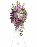 A large floral arrangement in a white three-legged stand, featuring purple and lavender flowers, white lilies, and green foliage, set against a plain white background.