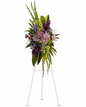 A large floral wreath on a white metal easel stand, featuring green foliage, purple and pink flowers, and white roses against a plain white background.